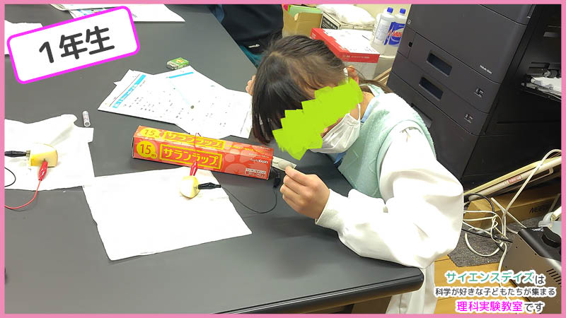 電子オルゴールに耳を近づけて音を確認する女の子 - 果物電池の成功を自分の耳で確かめるサイエンスデイズの授業