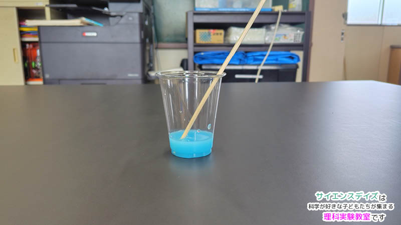 透明なカップに入れた水へ青い水性絵具を少量ずつ混ぜている様子