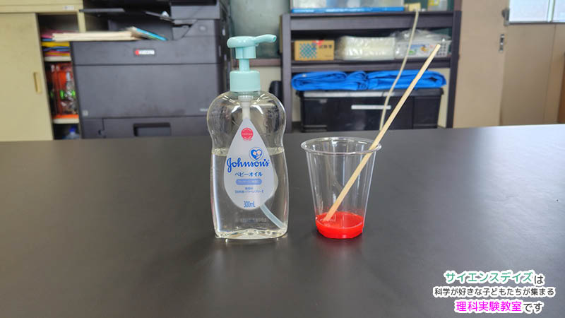 透明なカップのベビーオイルへ赤い油絵具を混ぜている様子